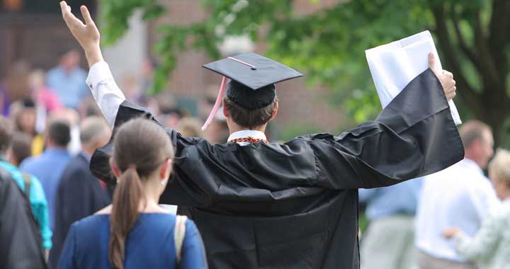 man in graduation cap holding hands up in air outside around other people
