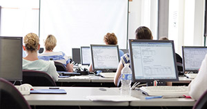 multiple monitor screens sitting on long desks with students at desks facing classroom whiteboard