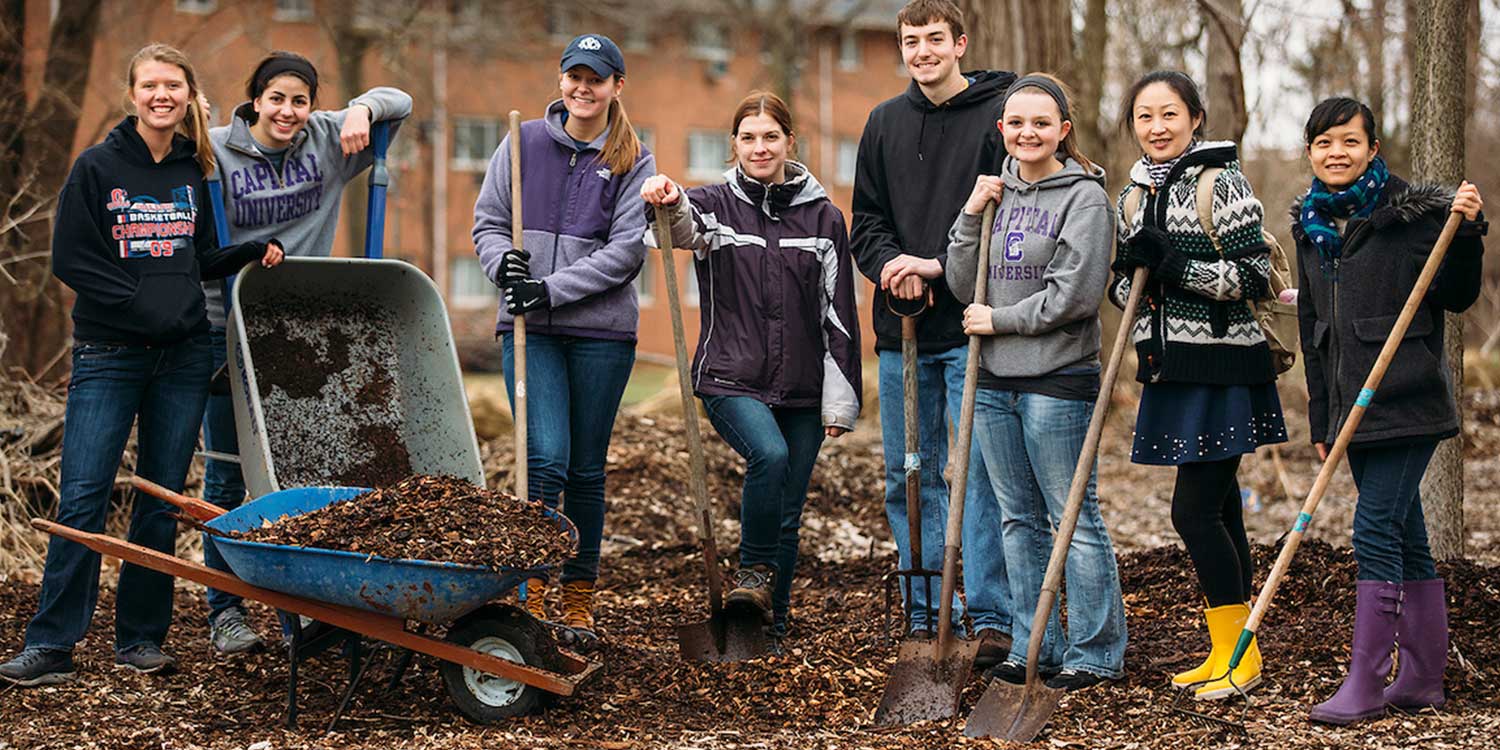 student community service team outside in mulch holding shovels and wheel barrows